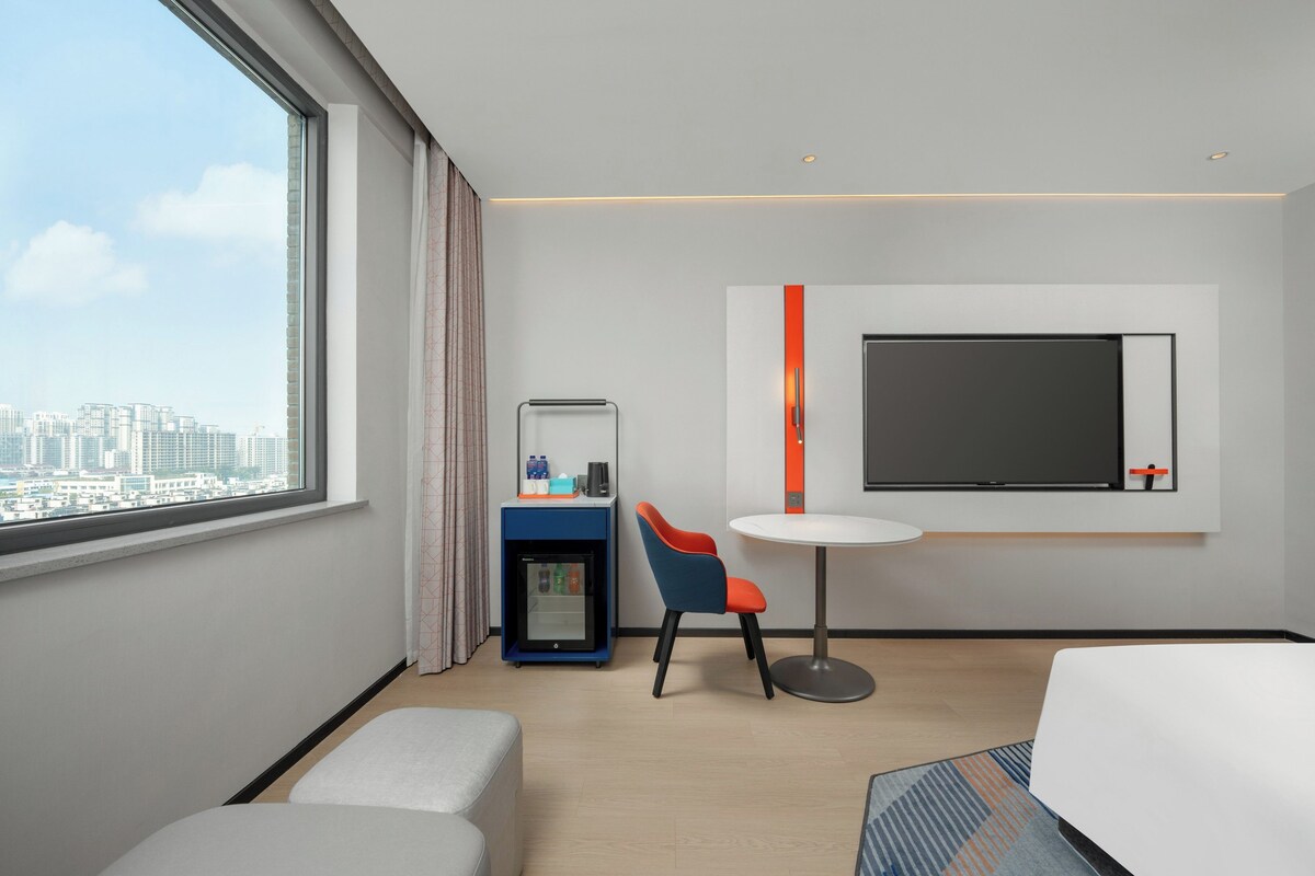 A modern hotel room with a large window, a flat-screen TV mounted on the wall, a small table, a blue and orange chair, a minibar, and a bed.