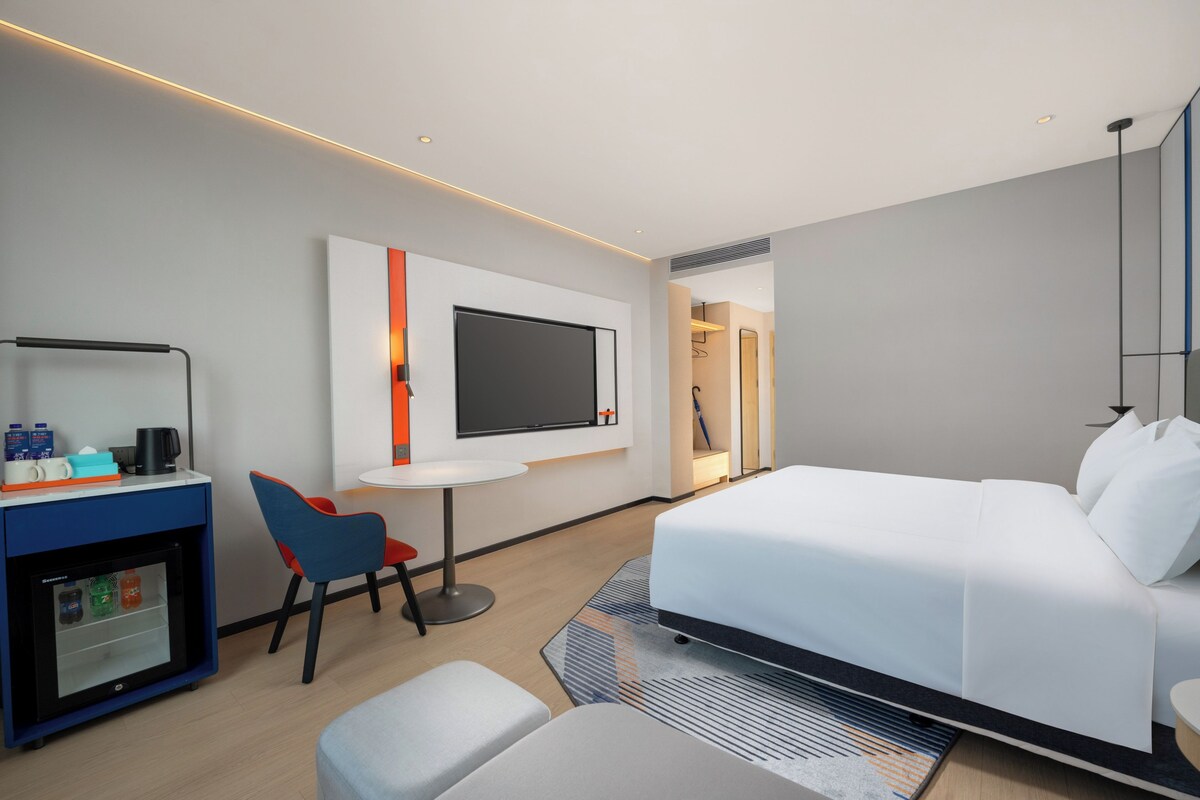 A modern hotel room with a bed, a desk with a computer, a small fridge, and a TV mounted on the wall.