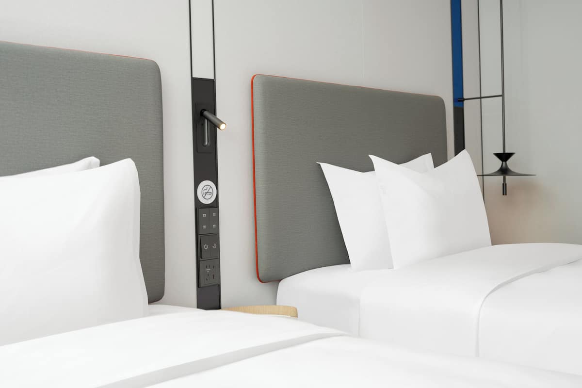 A hotel room with two beds, each with white bedding and grey headboards. There is a control panel with a power button and a light switch on the wall between the beds.
