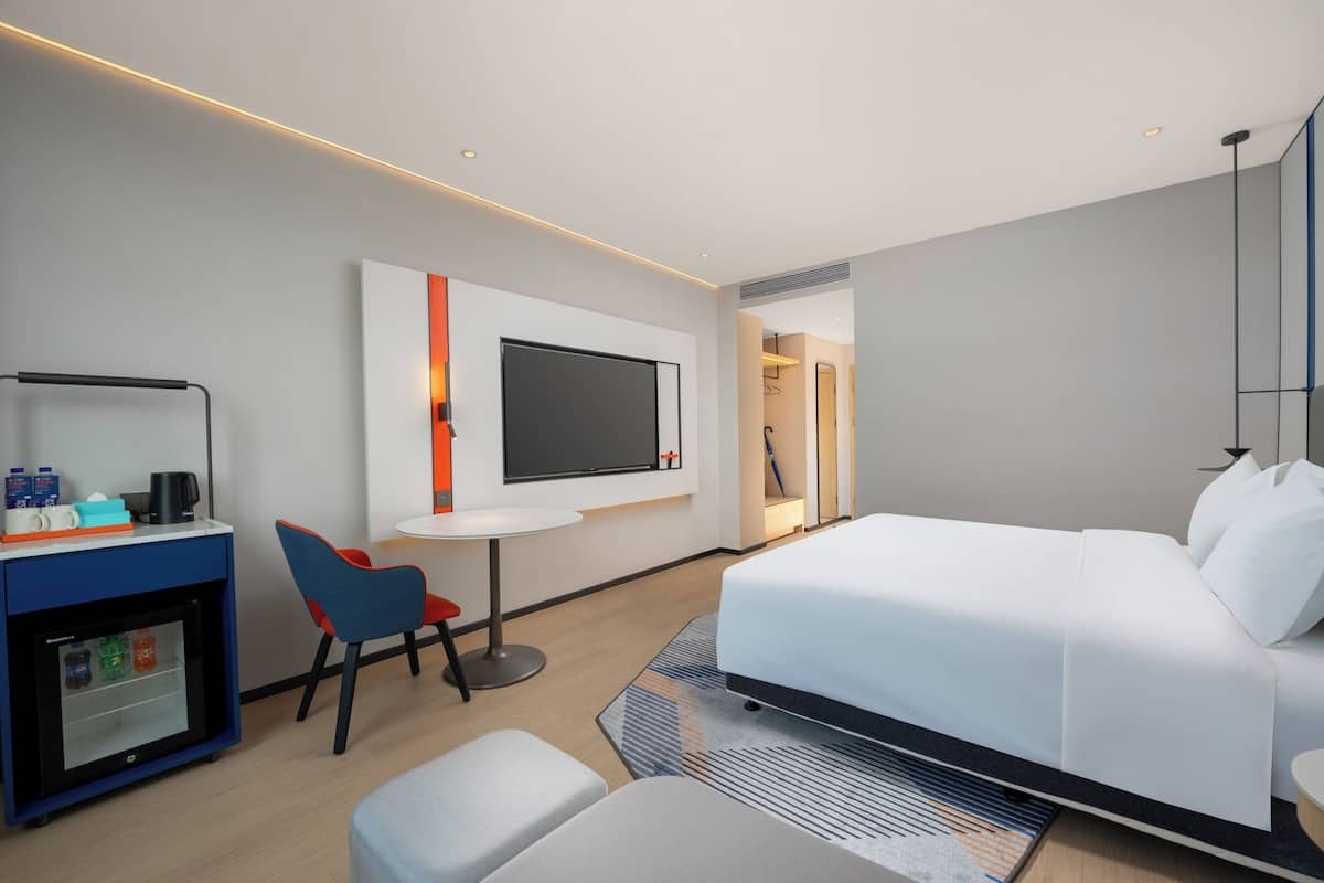 A modern hotel room with a bed, a desk with a computer, a small fridge, and a TV mounted on the wall.