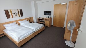 Una habitación de hotel con una cama de madera, un escritorio con televisor, un armario y un ventilador de pie.