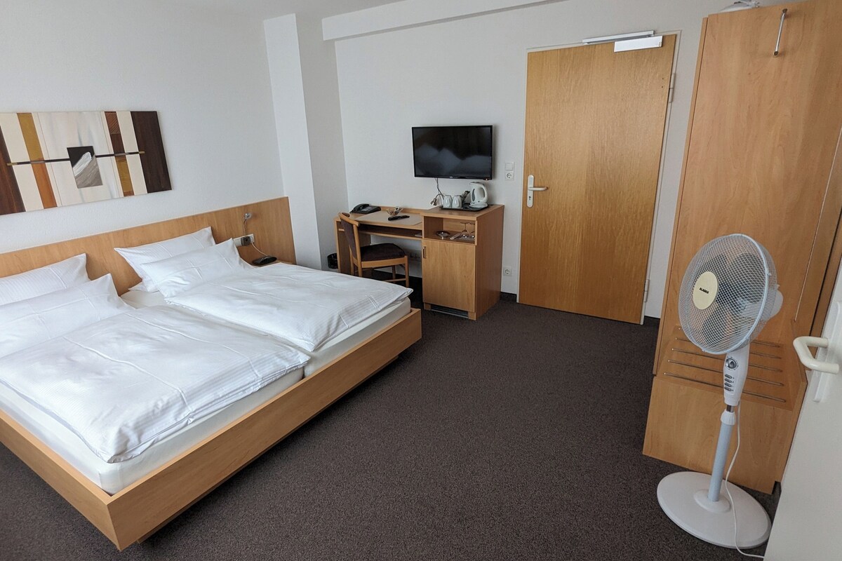 A hotel room with a wooden bed, a desk with a TV, a wardrobe, and a standing fan.