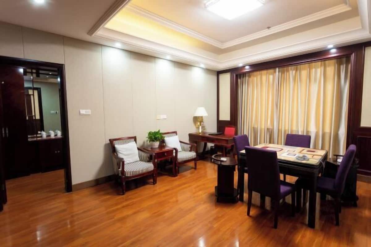 Une chambre d’hôtel avec un plancher en bois, une table à manger avec des chaises, un ensemble de canapés, une lampe et une grande fenêtre avec des rideaux.