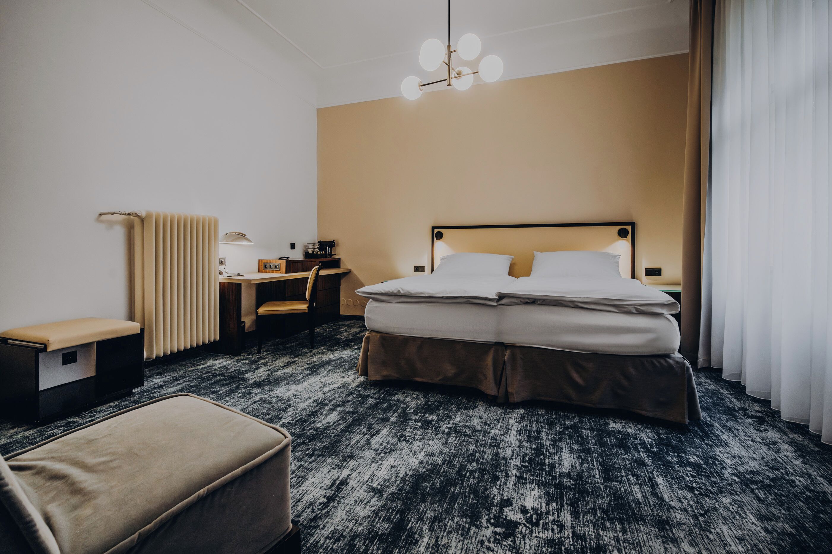 A hotel room with a large bed, a desk, a chair, a radiator, and a lamp.