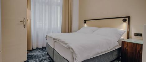 Una cama bien hecha con cabecera gris, una mesita de noche de madera con una lámpara y una ventana con cortinas translúcidas.