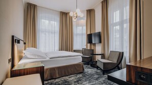 Habitación de hotel con cama, escritorio, silla, televisor y una ventana grande con cortinas.