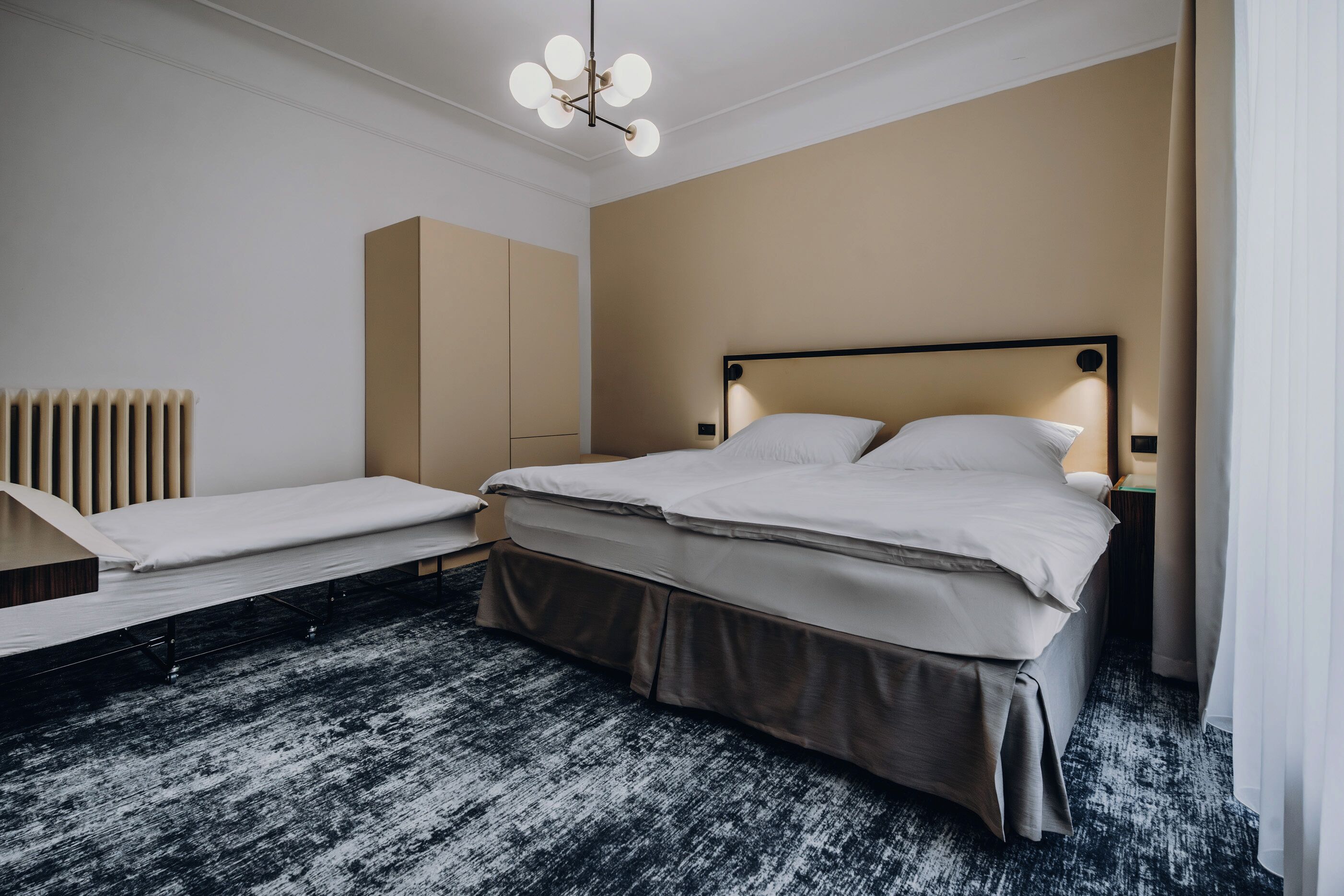 Quarto de hotel moderno com uma cama grande, uma cama menor, uma mesa de cabeceira, um abajur e um radiador.