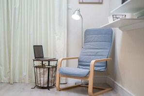 Una habitación con una silla tapizada de azul, una pequeña mesa redonda con una computadora portátil, una lámpara de pie y una cortina blanca.