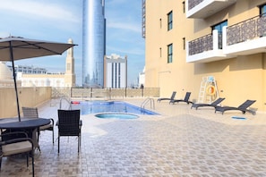 An outdoor pool area with lounge chairs and a table, a view of a tall skyscraper, and a building with balconies.