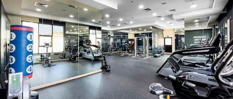 A modern gym with various exercise equipment, including treadmills and weight machines, and a mirrored wall.