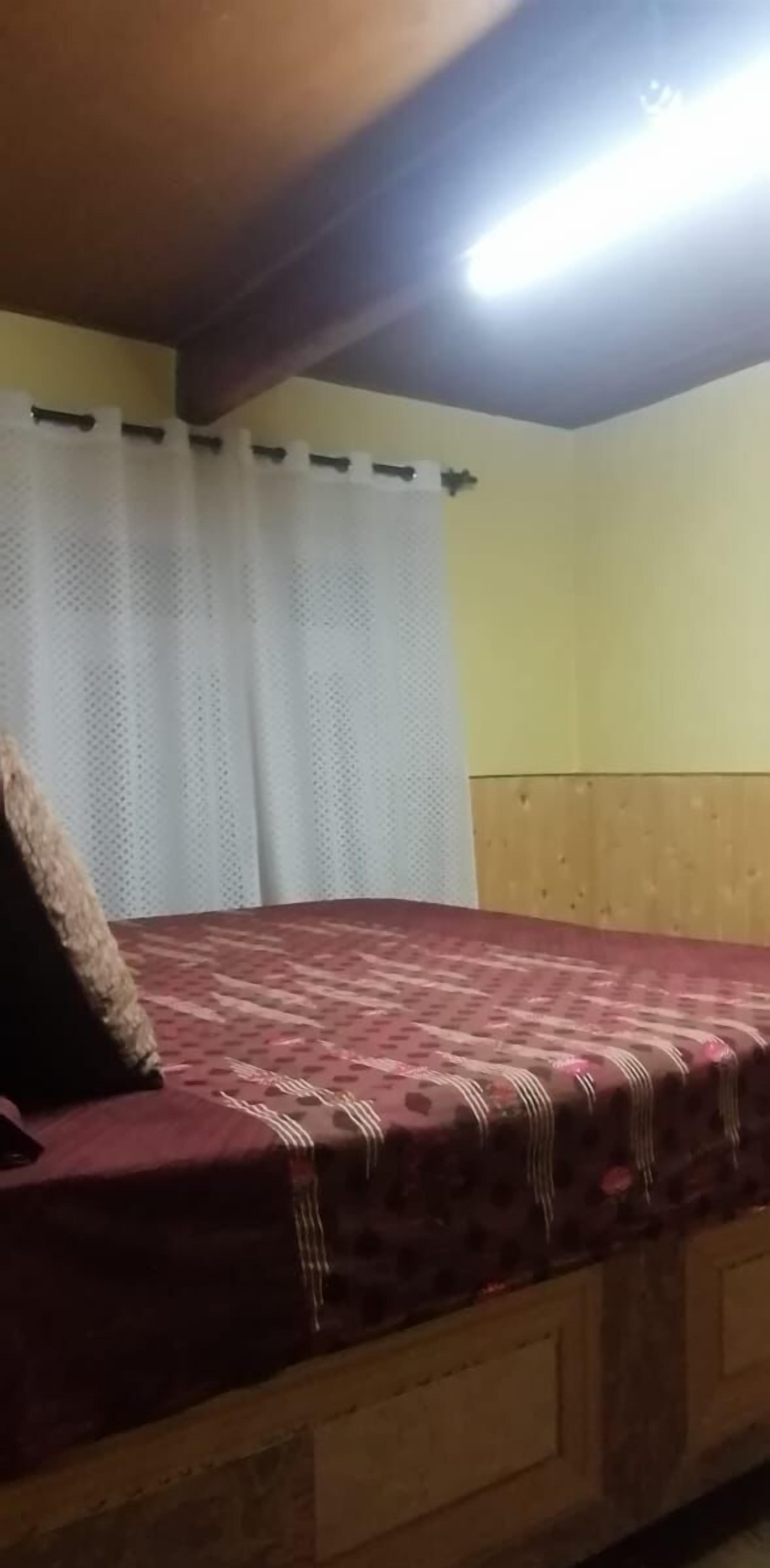 A single bed with a patterned bedspread in a room with wooden paneling and a white curtain.