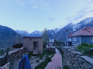 A narrow pathway leading to a small stone building with a door, surrounded by mountains and a house with a red roof. - Himalayaa Retreat (Lahul)