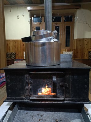 A wood stove with a pot on top, a fire burning inside, and a rustic interior with wooden walls and hanging star decorations. - Himalayaa Retreat (Lahul)