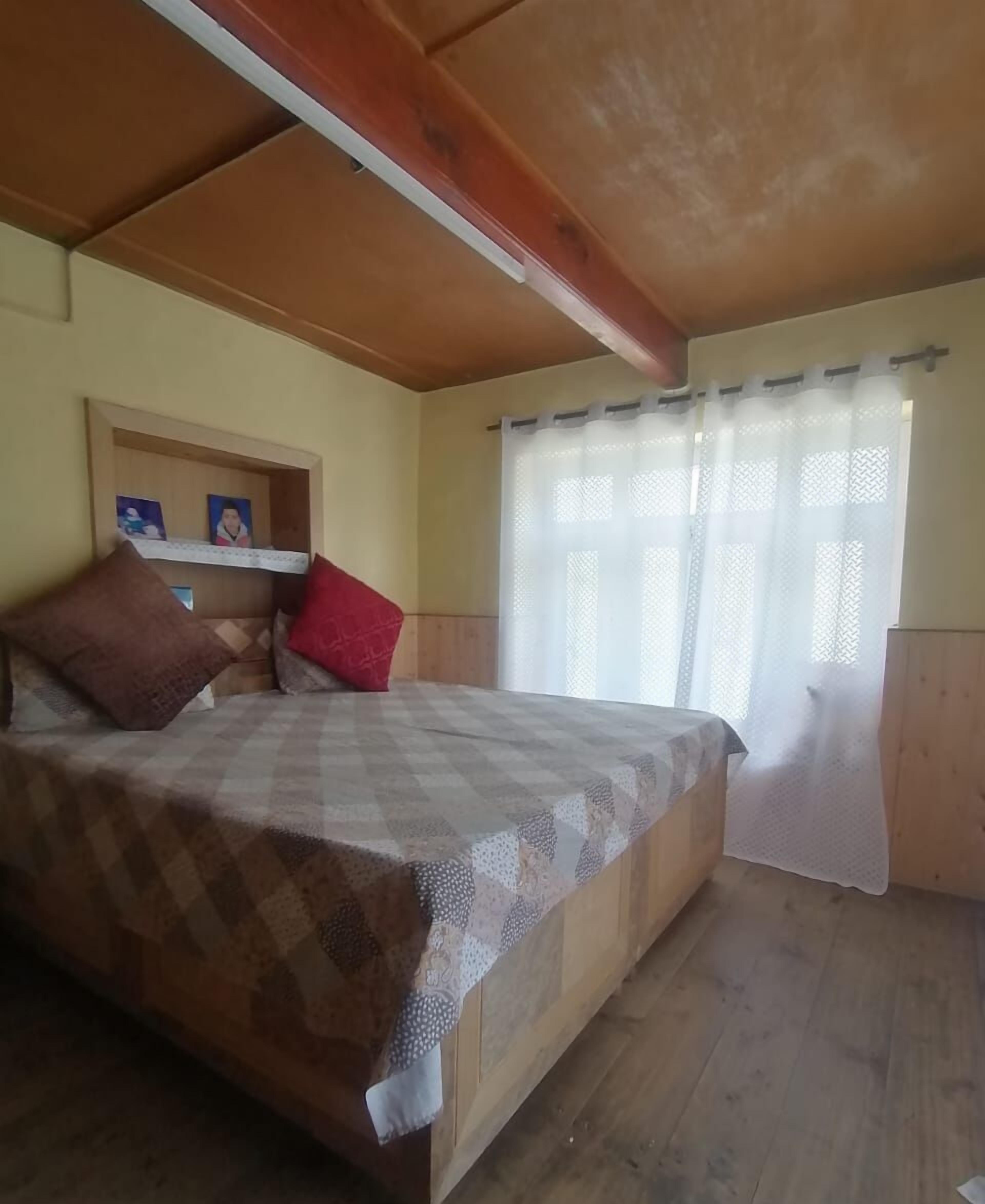 Ein Schlafzimmer mit einem hölzernen Bett, einer kleinen Ablage mit Bildern und einem Fenster mit transparenten Vorhängen.