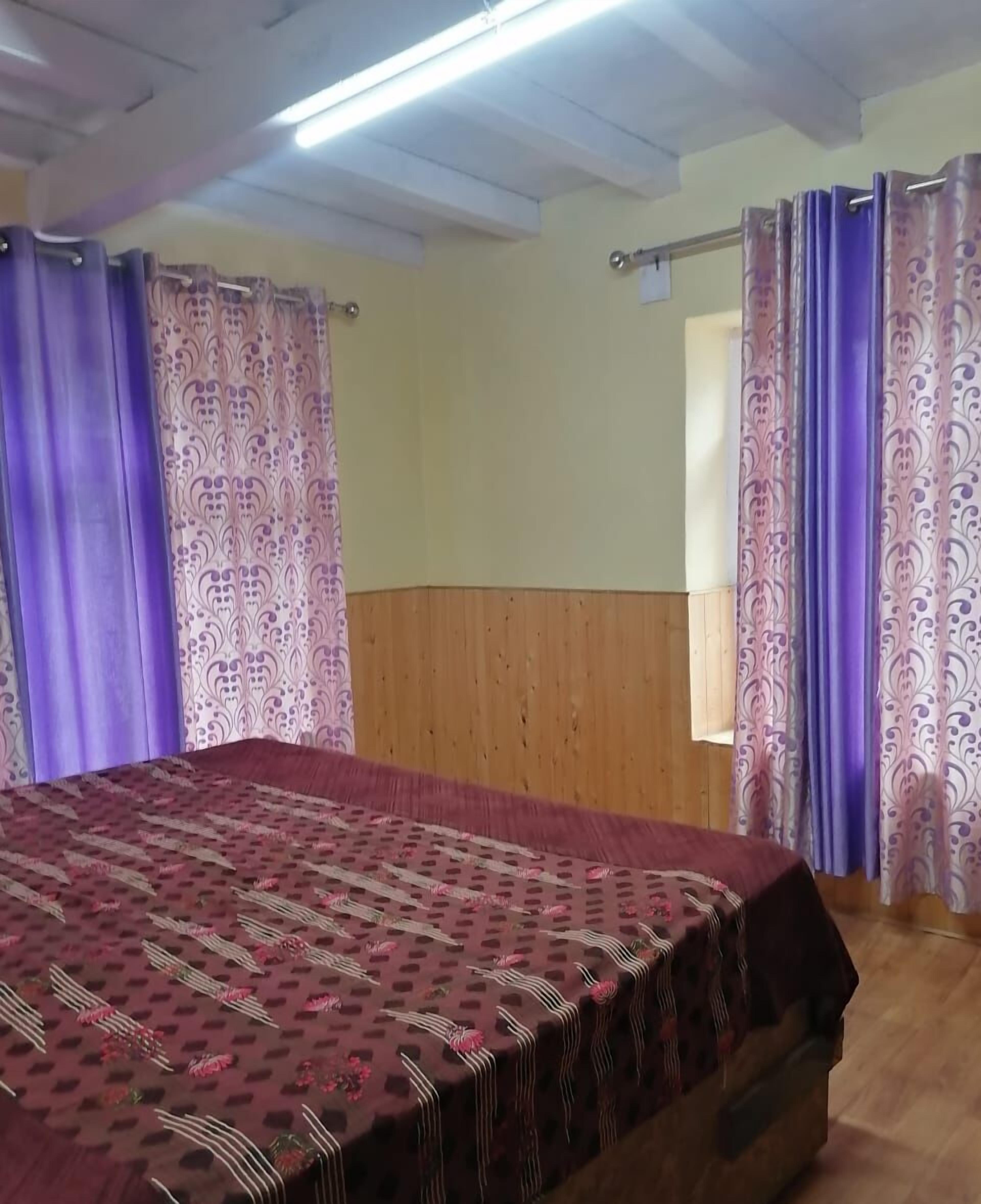 Une chambre dotée d’un grand lit, de lambris en bois aux murs, de rideaux violets et d’une couvre-lit à motifs.