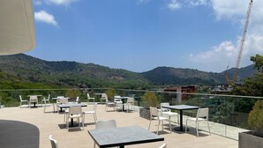 A rooftop terrace with tables and chairs, offering a view of a mountainous landscape and a construction site. - Mountainview Apartment in Naiharn (Rawai)
