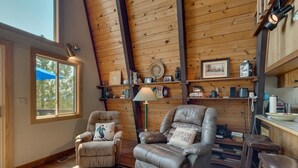 A cozy cabin interior with wooden walls, a large window, two leather recliners, a dining area, and a kitchen with shelves.