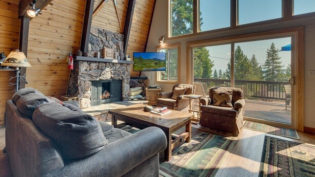 A cozy living room with a stone fireplace, wooden beams, and a large window overlooking a deck.