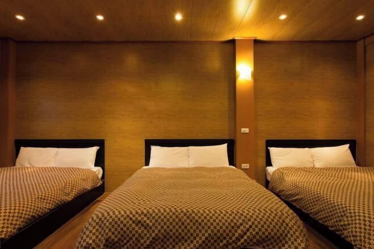 A hotel room with three single beds, each with a checkered bedspread and white pillows. The room has wooden walls and a wooden ceiling with recessed lighting.