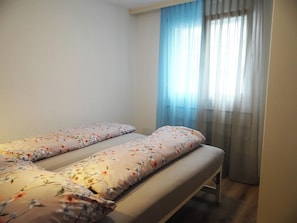 Habitación doble con dos camas individuales, una ventana con cortinas de encaje y una pared blanca lisa.