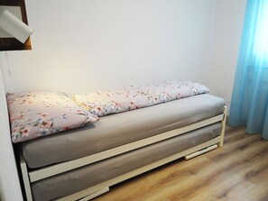 A single bed with a floral bedspread and a plain grey mattress in a room with wooden flooring and a light blue curtain.