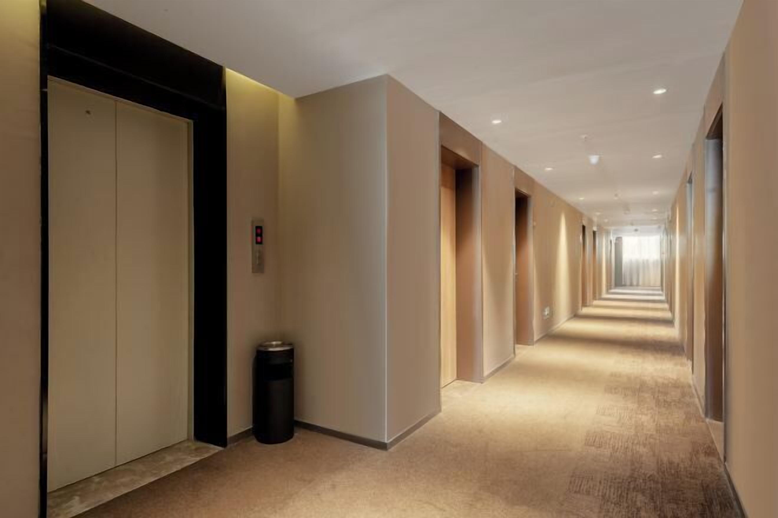 A long, narrow hotel corridor with beige walls, carpeted floors, and doors on the left.