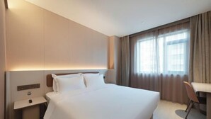 A hotel room with a large bed, bedside table, lamp, and a view of the outside through a window.