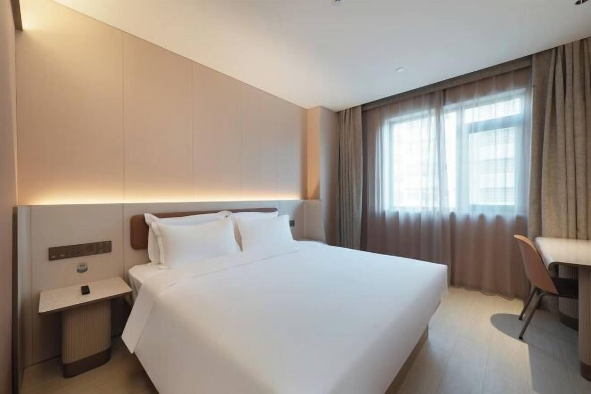 A hotel room with a large bed, bedside table, lamp, and a view of the outside through a window.