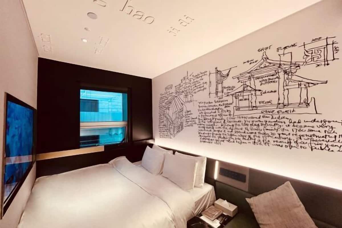 A modern hotel room with a large bed, a wall-mounted TV, and a large wall with architectural sketches.