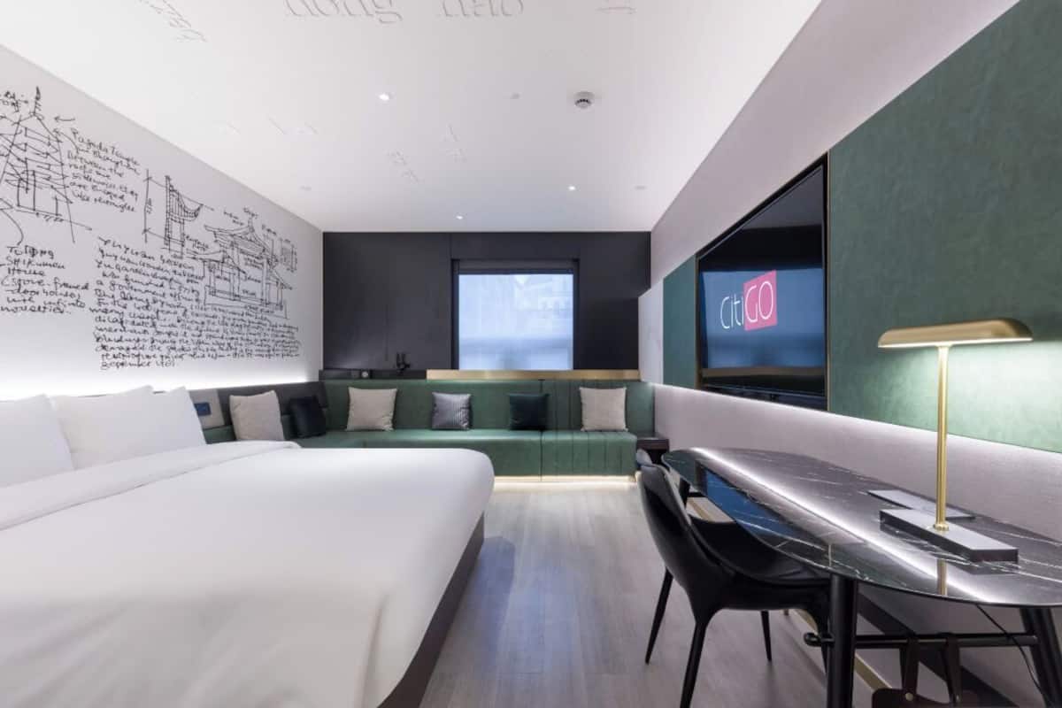 A modern hotel room with a bed, a desk, a chair, a large window, and a wall with architectural drawings.