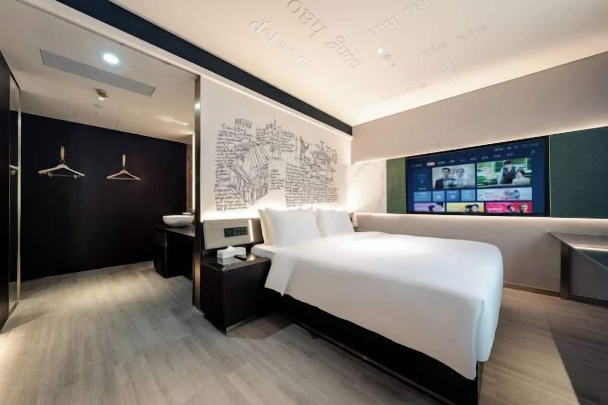 A modern hotel room with a large bed, bedside tables, a wall-mounted TV, and a cityscape artwork.
