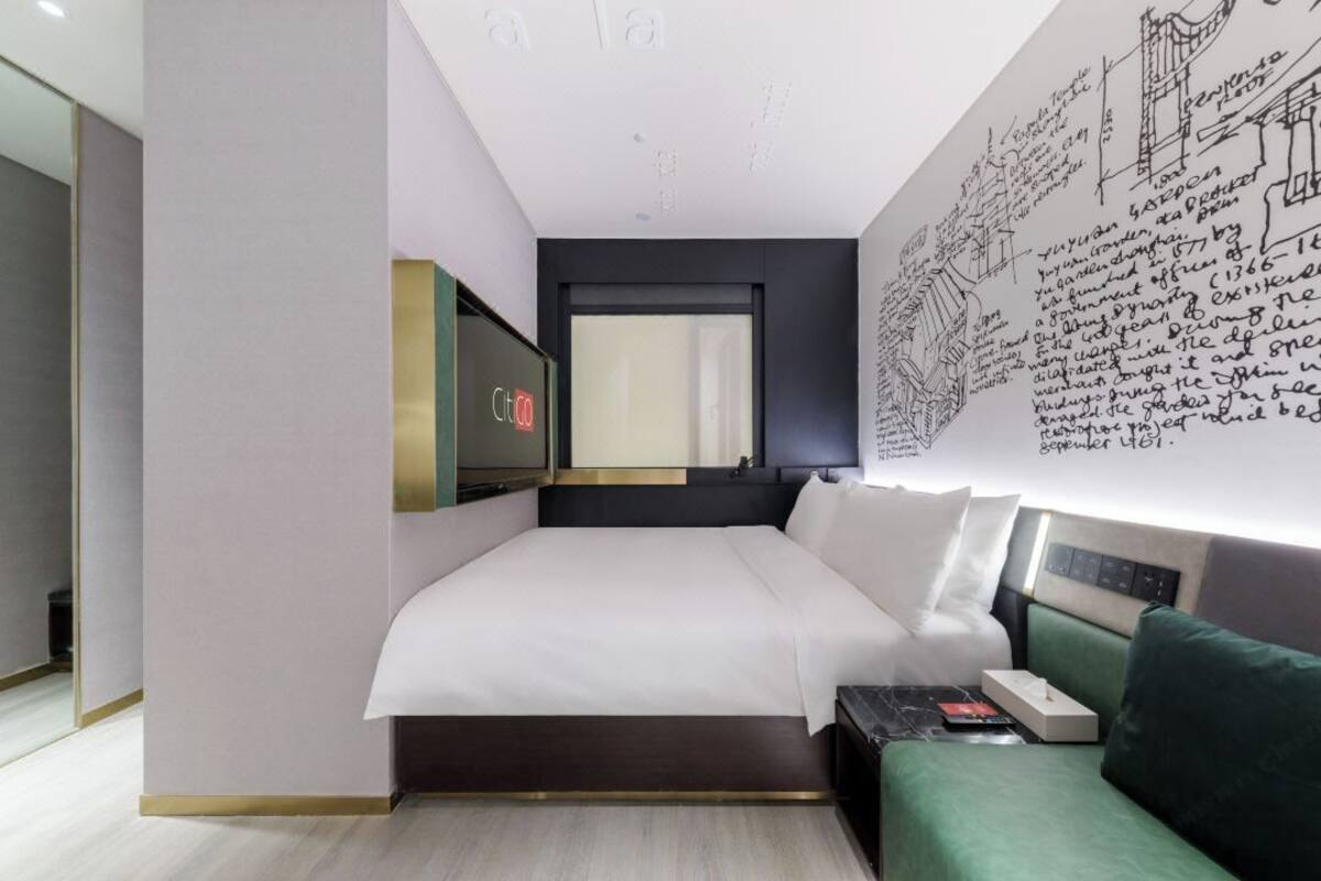 A modern hotel room with a large bed, a bedside table, a green sofa, and a wall with architectural sketches.