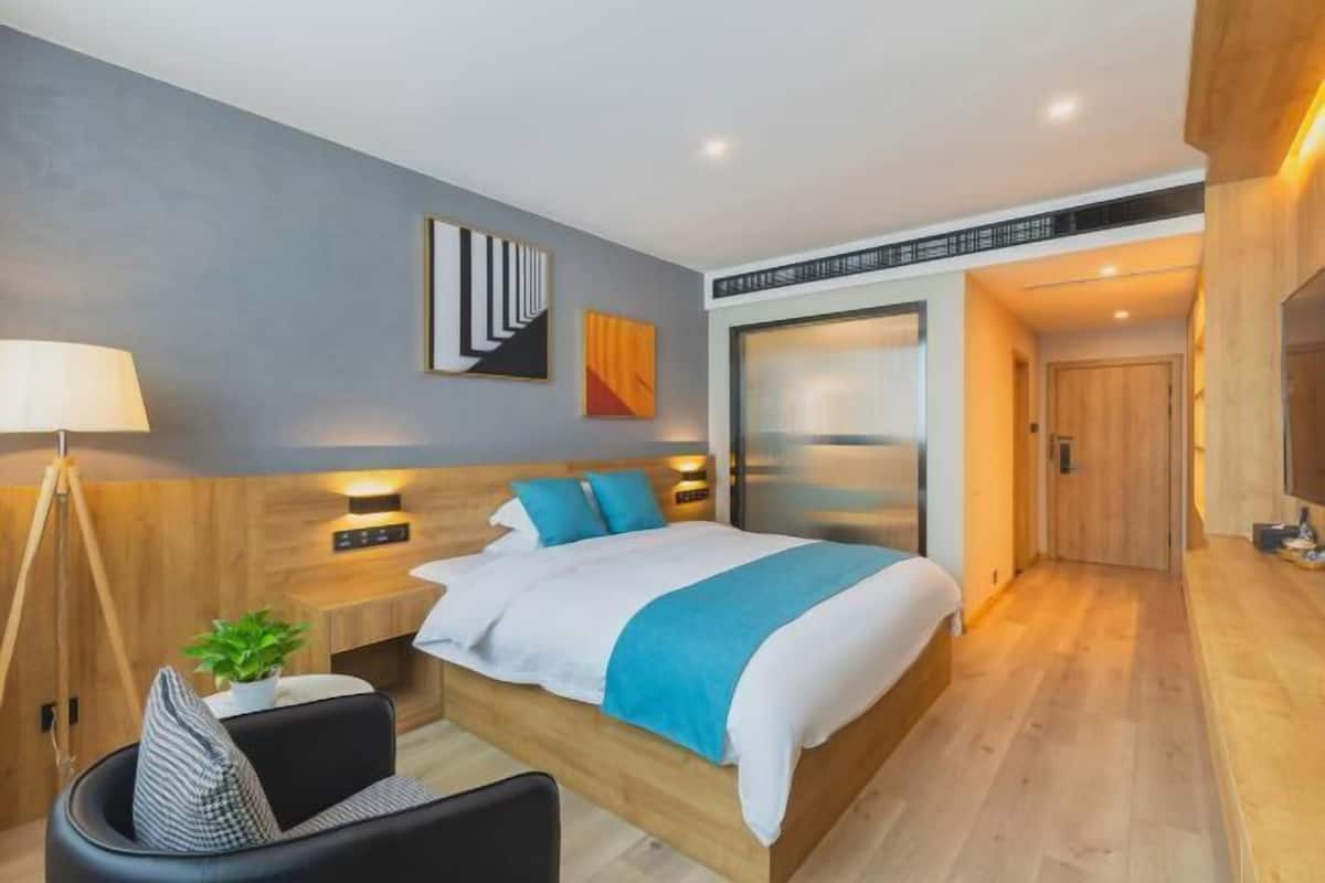 A modern hotel room with a large bed, bedside tables, a desk, a chair, a lamp, and a potted plant.