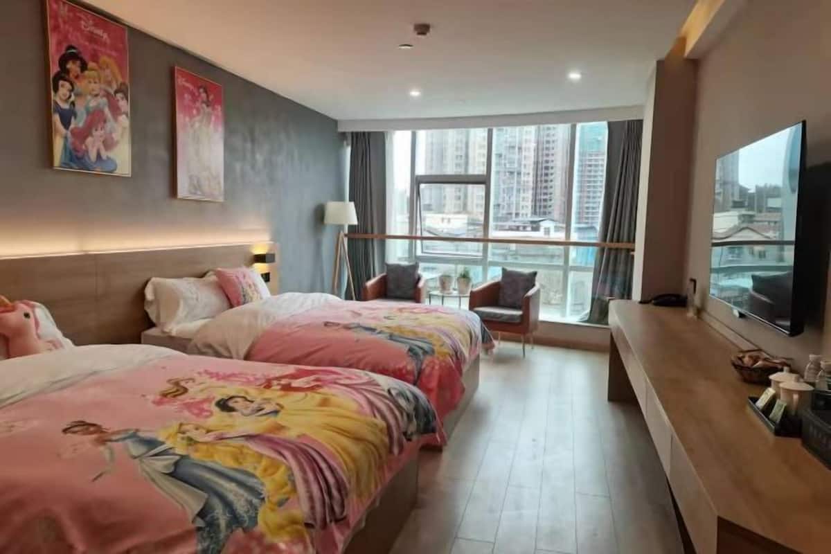 A hotel room with two beds, a TV, a desk, and large windows with a city view.