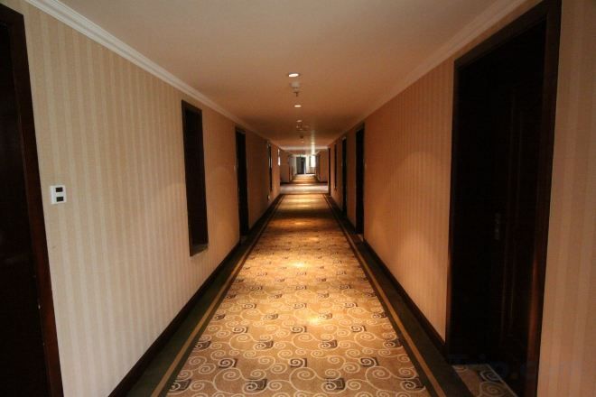 Un couloir d’hôtel avec un tapis à motifs, des portes en bois et un long couloir menant à une pièce plongée dans l’obscurité.