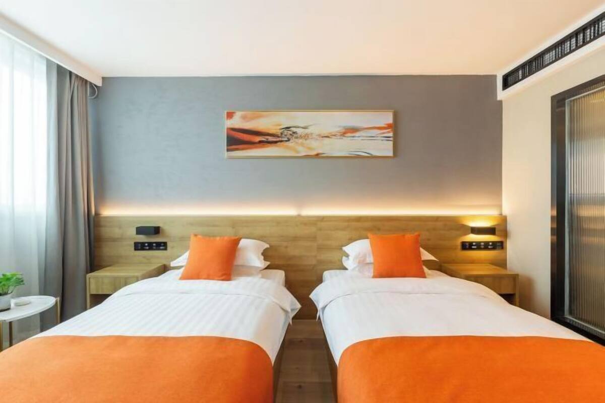 A hotel room with two beds, a painting on the wall, a nightstand with a lamp, and a window with curtains.