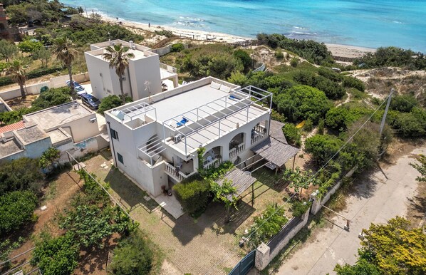 Um edifício moderno com telhado plano, rodeado por vegetação e uma praia ao fundo.