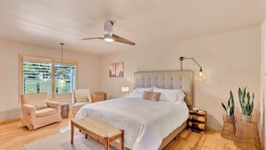 A bedroom with a large bed, a ceiling fan, a window with a view, a bedside table, and a bench.