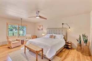 A bedroom with a large bed, a ceiling fan, a window with a view, a bedside table, and a bench.