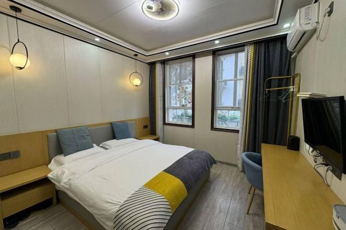 A hotel room with a bed, bedside table, chair, TV, and windows.