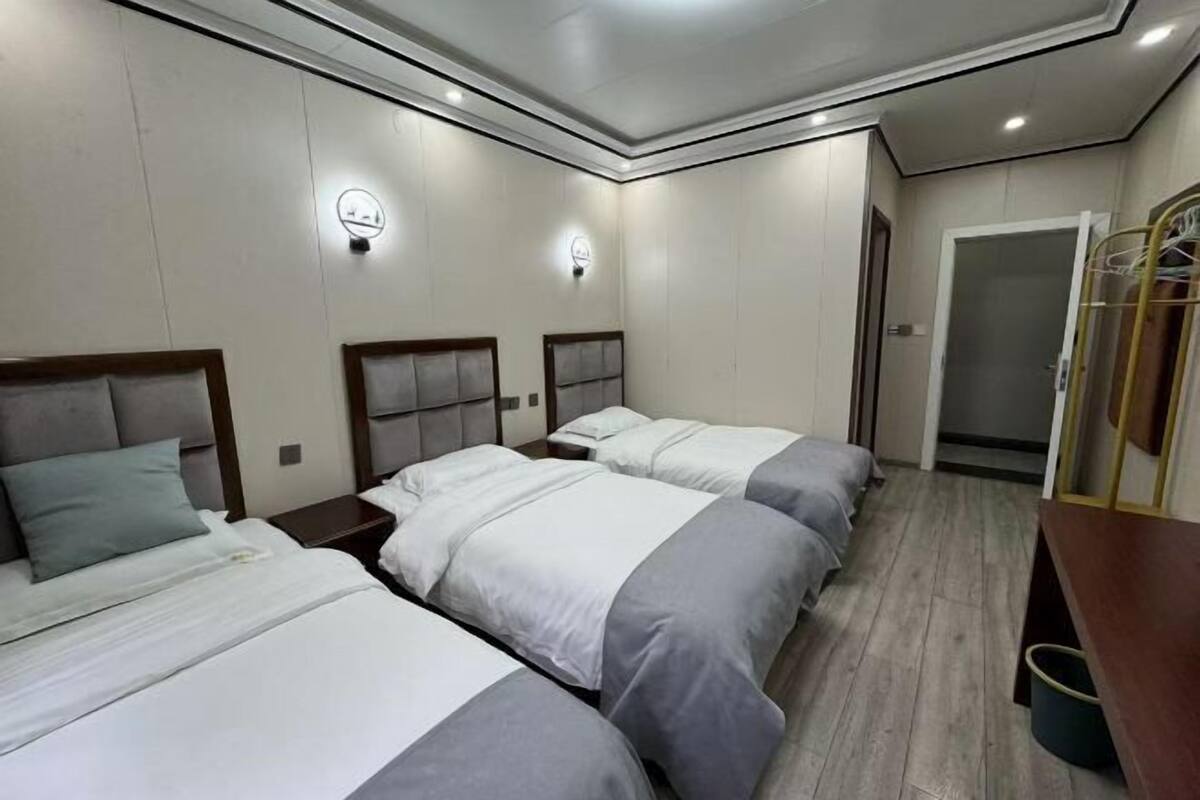 A hotel room with three beds, each with a wooden headboard and grey bedding. There are two wall-mounted light fixtures and a wooden bench with a trash bin.