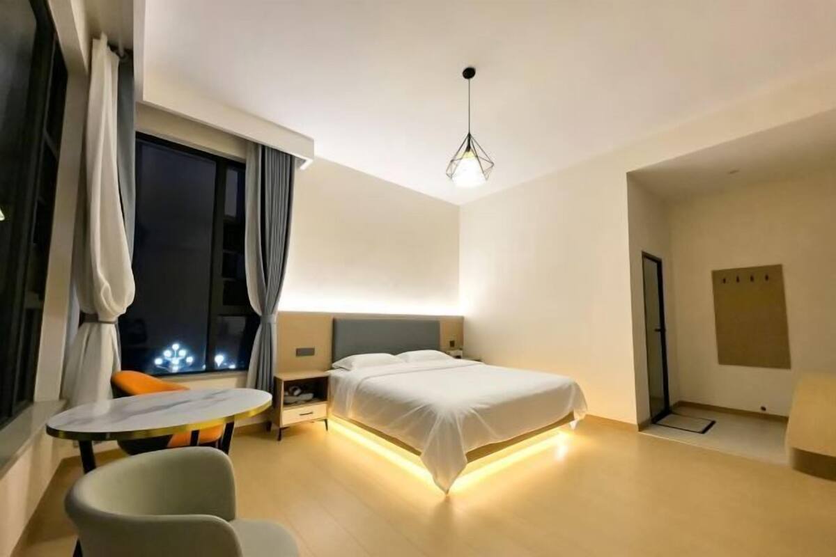 A modern hotel room with a bed, bedside table, lamp, and a view of the night sky through the window.
