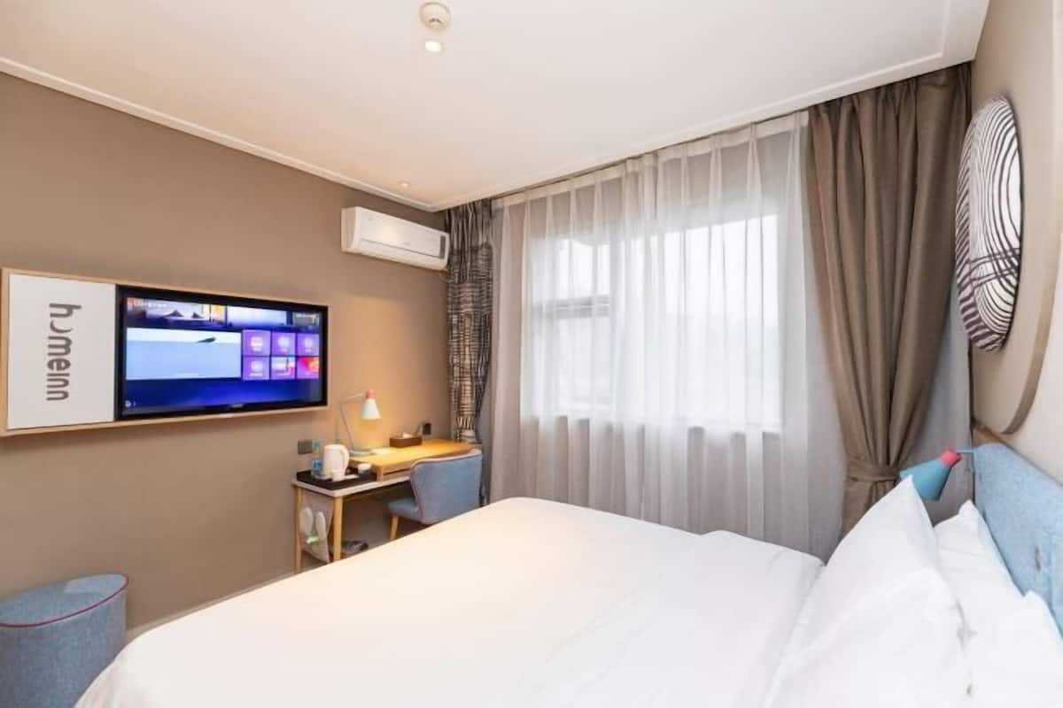 A hotel room with a bed, a desk with a chair, a TV mounted on the wall, and a window with curtains.