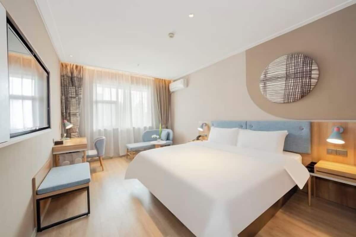 A hotel room with a large bed, a desk with a chair, a round wall decoration, and a bedside table with a lamp.