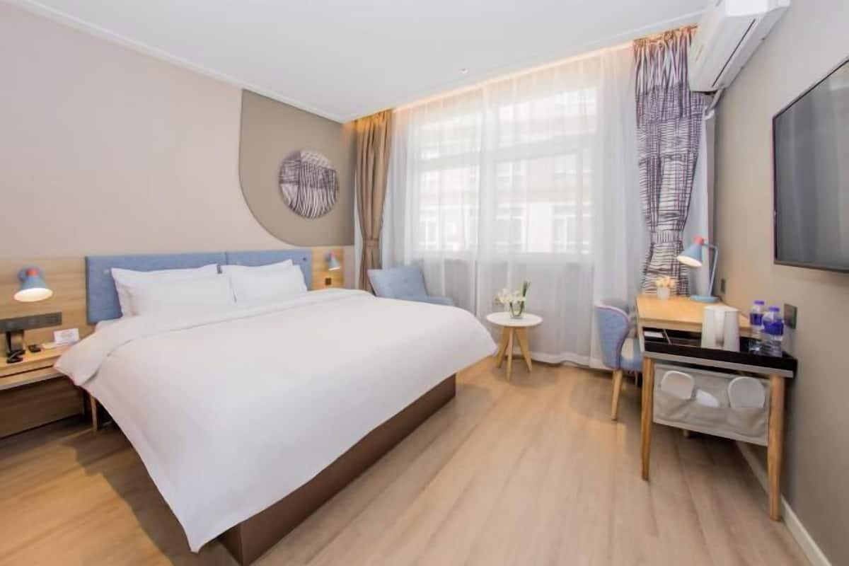 A hotel room with a large bed, a desk, a chair, a small table with a vase, and a TV mounted on the wall.