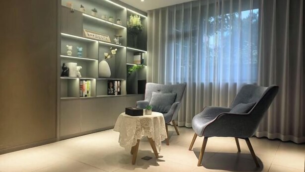A modern living room with a grey sofa, a small table with a lace tablecloth, and a bookshelf filled with decorative items.