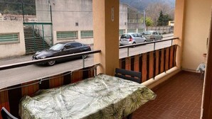 Un balcón con mesa y sillas, con vistas a una calle con coches aparcados y un edificio con escaleras.