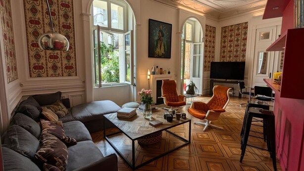 A spacious living room with a large sofa, a coffee table, and orange armchairs. There is a fireplace, a television, and large windows with patterned curtains. - Apartment with terrace, garden and WIFI in the historic heart of Angoulême (Angoulême)