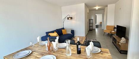 A modern living room with a dining area set for four. There is a sofa with cushions, a television, and a dining table with four chairs.
