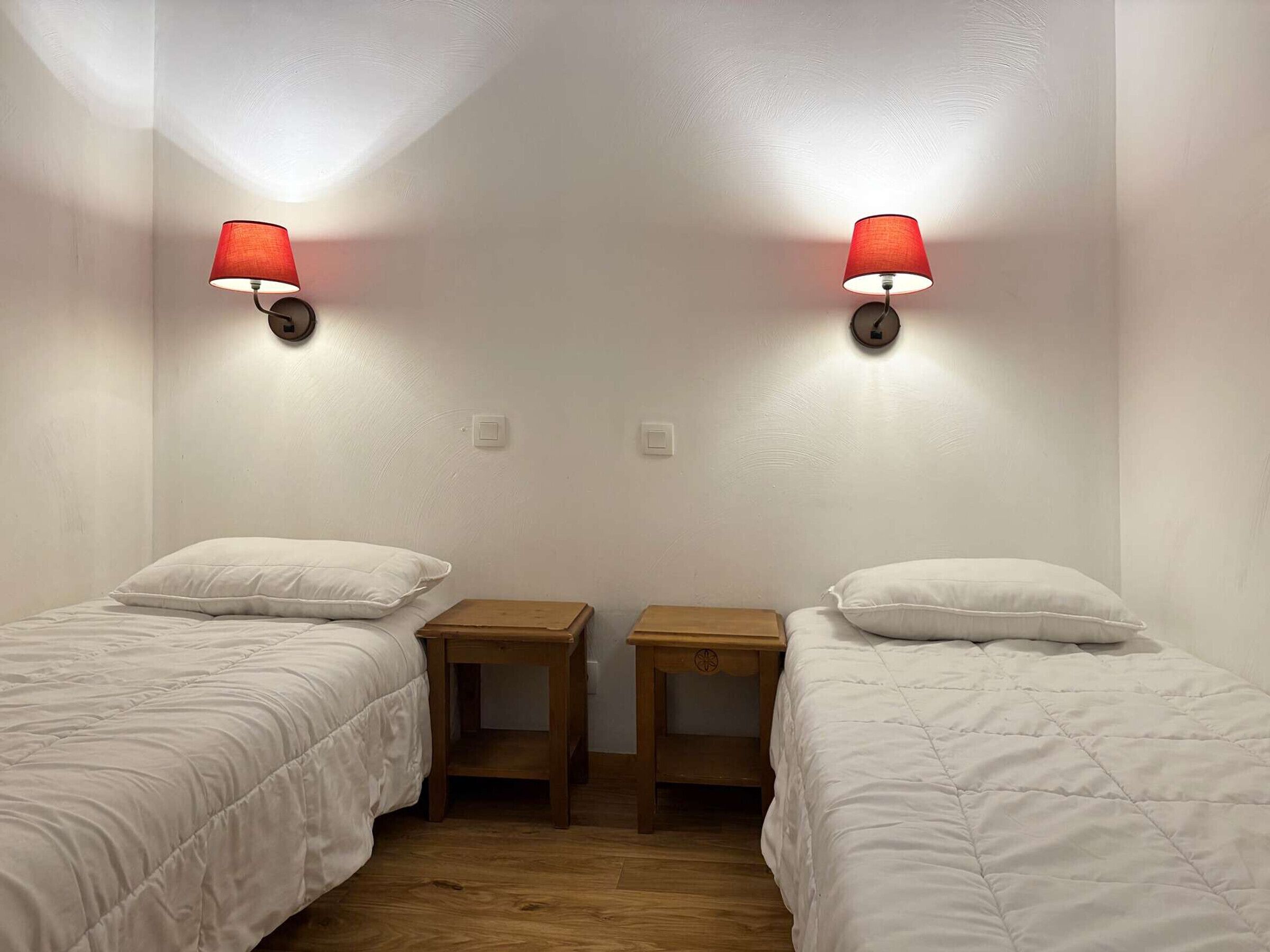 A room with two single beds, each with a white duvet and a wooden nightstand. There are two wall-mounted lamps with red shades. The room has white walls and a wooden floor.